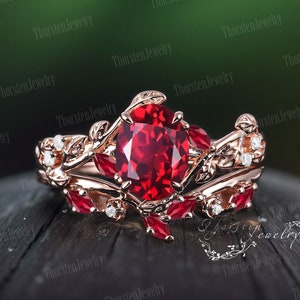 Könnte beinhalten: Ein Verlobungsring aus Roségold mit einem großen ovalen Rubin, der von kleineren Rubinen und Diamanten umgeben ist. Der Ring hat ein zartes florales Design mit Blättern und Ranken.