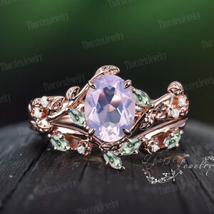 May include: A rose gold engagement ring set with a large oval-shaped pale pink gemstone surrounded by smaller clear gemstones. The ring band is decorated with delicate leaf and vine details. The matching wedding band features a row of clear gemstones.