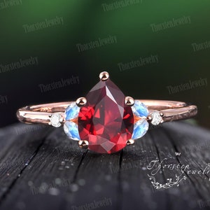 Könnte beinhalten: Ein Ring mit einem roten, birnenförmigen Edelstein und zwei kleineren blauen Edelsteinen auf jeder Seite. Der Ring ist in Roségold gefasst.