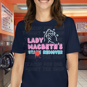 May include: Navy blue t-shirt with the text "LADY MACBETH'S STAIN REMOVER" in pink and blue, featuring a graphic of hands washing and a red stain. The shirt is from the "TEACUP FOR ONE T-SHIRT TEE SHOP".