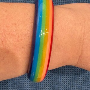May include: A smooth, rounded bangle bracelet featuring a vibrant, glossy rainbow pattern. The design consists of distinct, parallel vertical stripes in shades of purple, blue, green, yellow, orange, and red, wrapping around the entire circumference.