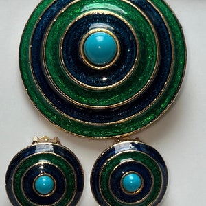 May include: A set of green, blue, and gold tone jewelry with turquoise accents. The set includes a brooch and a pair of earrings. The brooch is a large circle with concentric circles of green and blue enamel. The earrings are smaller circles with the same design.