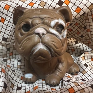 May include: A brown ceramic bulldog figurine with a white accent on its face and paws. The bulldog has a wrinkled face and is sitting down. The background is a patterned fabric with brown, orange, and white squares.
