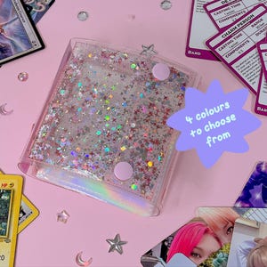 May include: A clear, glitter-filled card holder with pink accents and a star-shaped clasp. The holder is surrounded by trading cards and decorative elements on a pink surface. Text on a purple starburst reads "4 colours to choose from."