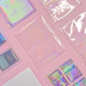 Può includere: Una collezione di buste trasparenti per raccoglitori con effetto olografico iridescente disposte su una superficie rosa. I fogli in plastica presentano diverse fessure per organizzare carte collezionabili o inserti in carta.