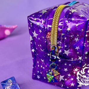 May include: A clear purple zippered pouch with a galaxy print and silver stars. The pouch has a silver metal charm attached to the zipper pull, shaped like a multi-colored dice. The pouch has the text "Imaginary Friends" printed on the bottom.