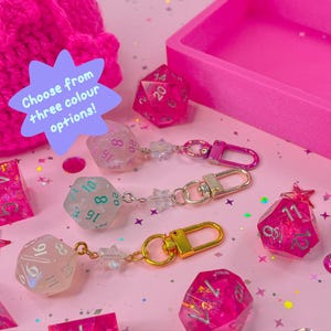 May include: A selection of pink dice keyrings with star charms and gold or silver clasps. The image features different dice shapes and sizes, with the text "Choose from three colour options!"