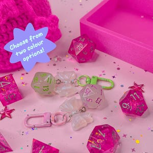 Puede incluir: Una colección de dados rosas y transparentes, llaveros y un recipiente rosa sobre una superficie rosa. Los dados tienen varias formas, incluido un dado de 20 caras. Algunos dados están unidos a llaveros con cierres rosas y verdes. El texto en una estrella dice "¡Elige entre dos opciones de color!"