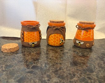 May include: Three decorative jars with orange and yellow honeycomb patterns, brown rope accents, and bee embellishments. Each jar has a cork lid. One lid is separate. The jars are on a dark surface.