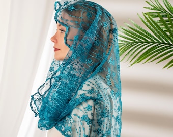 May include: A teal lace veil with floral embroidery. The veil is draped over a person's head and shoulders, with the intricate lace design visible. The background includes a white curtain and a green plant.