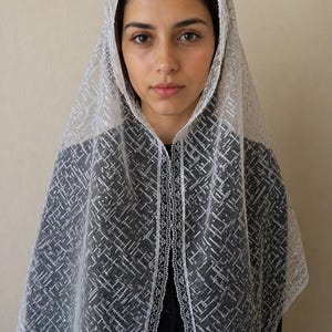 May include: A white lace veil with a geometric pattern, draped over the head and shoulders. The veil has a delicate, open weave design and a scalloped edge. The person is wearing a black top.
