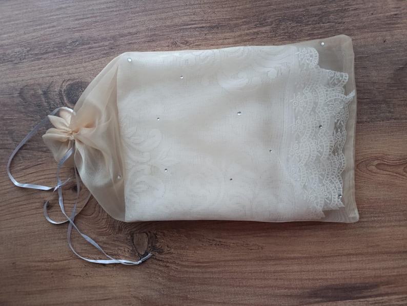 May include: A white sheer fabric bag with a lace trim and small rhinestones. The bag is tied with a white ribbon.