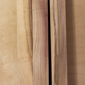 May include: Three unfinished wooden planks with natural wood grain. The planks vary in width and feature light brown and reddish-brown hues. The wood appears to be ready for use in a woodworking project.