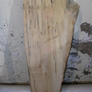 May include: A light-colored, unfinished wooden slab with a natural edge. The wood grain features dark, linear markings and knots. Pink markings are visible on the surface. The slab is supported by a small wooden base. Ideal for woodworking projects.