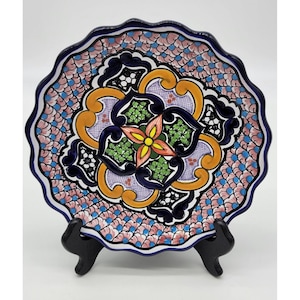 May include: A decorative ceramic plate with a scalloped edge. The plate features a colorful floral design with blue, green, yellow, orange, and pink accents. The design is centered on a large flower with four petals.
