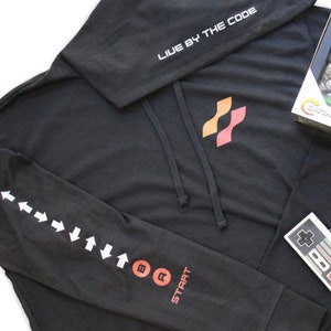May include: Black hooded sweatshirt with a red and orange logo on the front and the text "LIVE BY THE CODE" on the hood. The left sleeve has white arrows and red buttons labeled "START", "A", and "B".