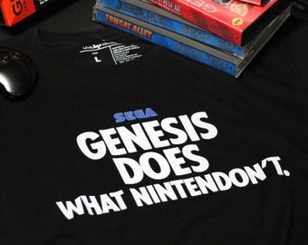 May include: A black t-shirt with white text that reads "SEGA GENESIS DOES WHAT NINTENDO DON'T."  The shirt is laid out on a table with a stack of Sega Genesis game cartridges.