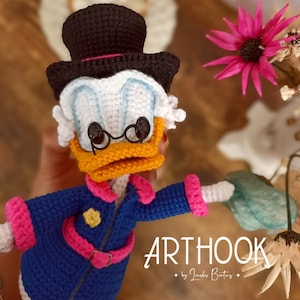 Puede incluir: Juguete de amigurumi tejido a crochet de Donald Duck con un sombrero de copa negro, una camisa blanca, una chaqueta azul y detalles rosas. El juguete tiene un pico amarillo, pies naranjas y ojos negros. El juguete sostiene una flor verde tejida a crochet. El texto "ARTHOOK by Jordeis Beatriz" es visible en la imagen.