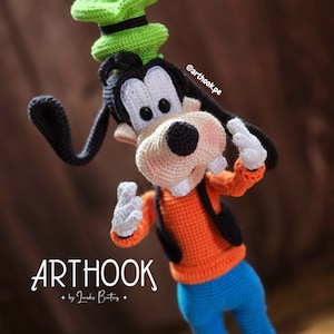 Könnte beinhalten: Gehäkeltes Amigurumi-Spielzeug von Goofy von Disney, das einen grünen Hut und ein orangefarbenes Hemd mit einer schwarzen Weste trägt. Das Spielzeug steht mit erhobenen Armen und hat einen freundlichen Ausdruck. Der Text "ARTHOOK by Janis Butirs" ist unter dem Spielzeug sichtbar.