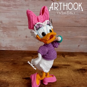 Può includere: Una bambola amigurumi all'uncinetto di un'anatra dei cartoni animati che indossa un maglione viola e un fiocco rosa. La bambola è in piedi su una superficie di legno. Il testo "ARTHOOK by Jandres Beatriz" è visibile sopra la bambola.