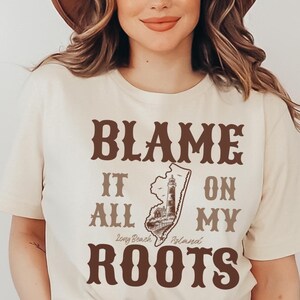 May include: A cream-colored short-sleeve t-shirt featuring brown text that reads BLAME IT ON MY ROOTS, with a graphic of the state of New Jersey containing a lighthouse and the text Long Beach Island, paired with a brown wide-brimmed hat.