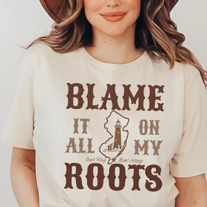May include: A cream-colored short-sleeve t-shirt featuring brown text that reads BLAME IT ON MY ROOTS. The design includes a map outline of New Jersey with a lighthouse graphic and the text Cape May New Jersey centered within the shirt design.