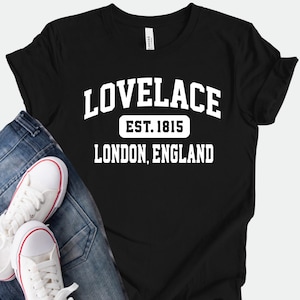 Könnte beinhalten: Schwarzes T-Shirt mit dem weißen Text "LOVELACE EST. 1815 LONDON, ENGLAND". Dazu eine blaue Jeans und weiße Turnschuhe mit roten Akzenten.