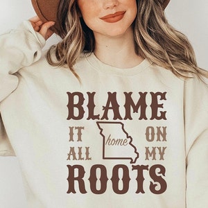 May include: A cream-colored crewneck sweatshirt featuring brown text that reads BLAME IT ALL ON MY ROOTS with a central outline of the state of Missouri containing the word home. The garment is paired with a wide-brimmed brown felt hat.