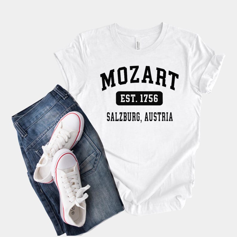 May include: White t-shirt with black text that reads "Mozart Est. 1756 Salzburg, Austria". The shirt is paired with blue jeans and white sneakers.
