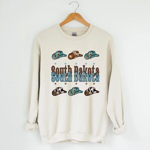 Peut inclure: Sweat-shirt crème avec l'inscription "South Dakota" en marron et turquoise, orné de six chapeaux de cow-boy aux motifs variés. Les chapeaux sont dans des tons de marron, turquoise et noir et blanc. Le design comprend de petites étoiles.