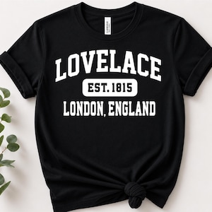 May include: A black short-sleeved t-shirt featuring white collegiate-style lettering that reads LOVELACE, EST. 1815, and LONDON, ENGLAND. The shirt has a crew neckline and a knotted hem detail at the bottom, with a small brand tag inside the collar.