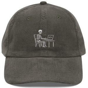 May include: A dark gray corduroy baseball cap featuring an embroidered white graphic of a skeleton sitting at a desk on a chair, typing on a laptop that displays the code symbol < /> on its screen, centered on the front panel of the hat.