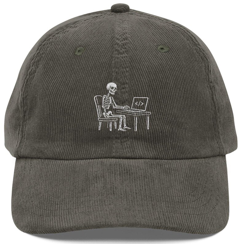 May include: A dark gray corduroy baseball cap featuring an embroidered white graphic of a skeleton sitting at a desk on a chair, typing on a laptop that displays the code symbol < /> on its screen, centered on the front panel of the hat.