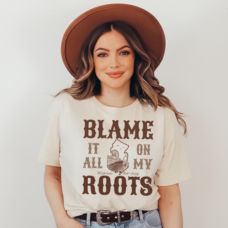 Op de afbeelding: Een cr&egrave;mekleurig T-shirt met korte mouwen en bruine tekst die luidt BLAME IT ALL ON MY ROOTS, met een centrale afbeelding van een reuzenrad en een promenade, en de tekst Wildwood New Jersey onder de afbeelding, gecombineerd met een bruine hoed.