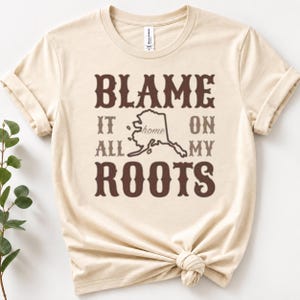 以下が含まれることがあります： クリーム色の半袖Tシャツ。ダークブラウンの文字で「BLAME IT ALL ON MY ROOTS」とプリントされ、中央にはアラスカ州の輪郭が描かれています。その中には、より明るいブラウンの筆記体で「home」という文字が入っています。