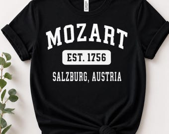 May include: A black short-sleeved t-shirt featuring white collegiate-style text that reads MOZART, followed by a white rectangular box containing the black text EST. 1756, and the text SALZBURG, AUSTRIA printed in white below the box.
