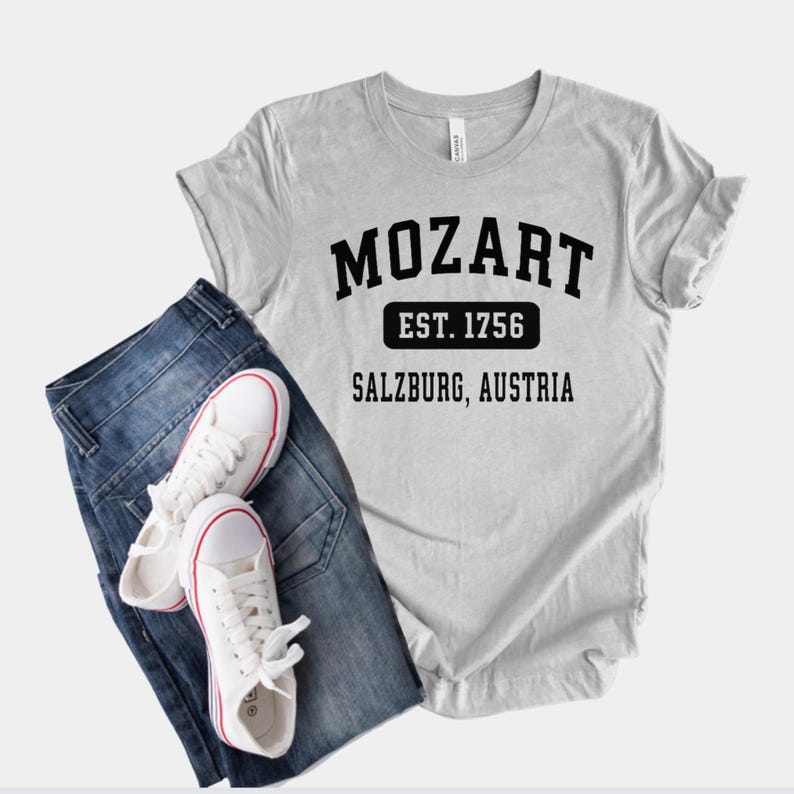 May include: A gray t-shirt with black text that reads "Mozart Est. 1756 Salzburg, Austria". The shirt is paired with blue jeans and white sneakers.