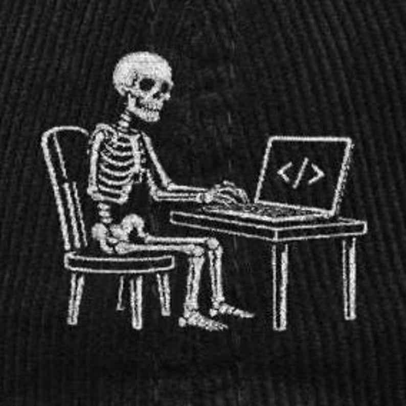 May include: A white embroidered graphic on a black fabric background depicting a human skeleton sitting on a chair at a desk. The skeleton is typing on a laptop computer that displays the code symbol </> on its screen in a simple white line art style.