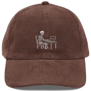 May include: A brown corduroy baseball cap featuring a white embroidered graphic of a skeleton sitting on a chair at a desk while working on a laptop. The laptop screen displays the code symbol consisting of a slash between two angle brackets.
