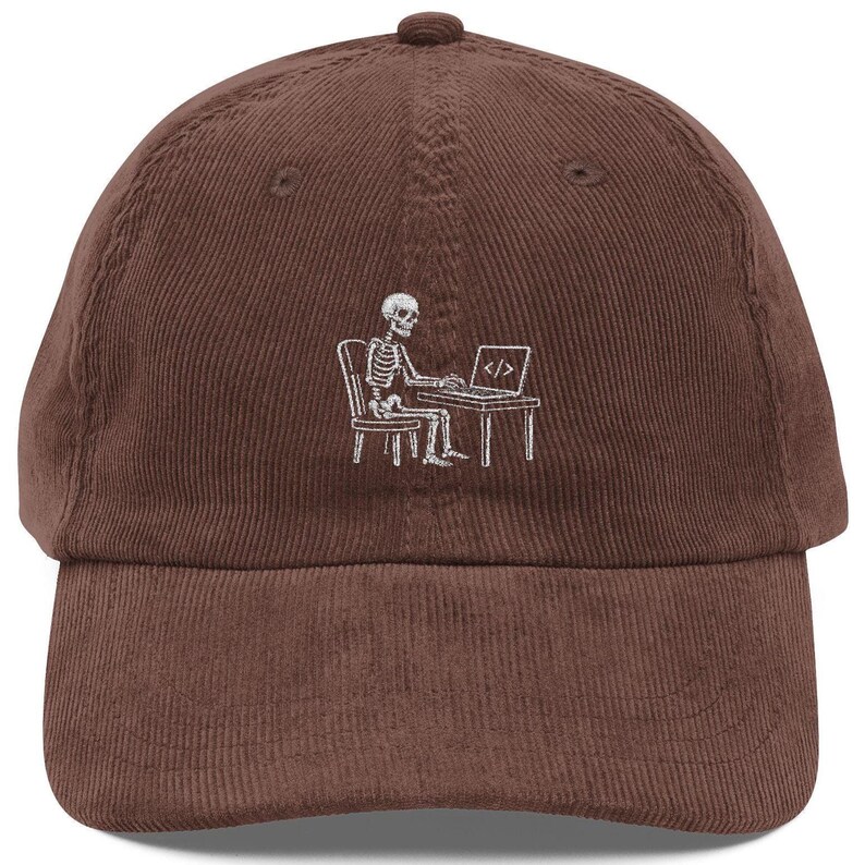 May include: A brown corduroy baseball cap featuring a white embroidered graphic of a skeleton sitting on a chair at a desk while working on a laptop. The laptop screen displays the code symbol consisting of a slash between two angle brackets.