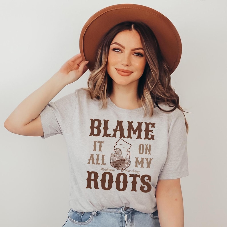 Op de afbeelding: Een lichtgrijs T-shirt met korte mouwen en bruine tekst BLAME IT ON ALL MY ROOTS met een afbeelding van de staat New Jersey en een reuzenrad. Het shirt wordt gecombineerd met een bruine hoed met brede rand en een lichtblauwe spijkerbroek.