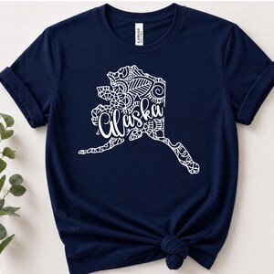 以下が含まれることがあります： アラスカ州の白いグラフィックが描かれたネイビーブルーの半袖Tシャツ。地図の輪郭は複雑なマンダラ風の模様で埋め尽くされており、中央には白い筆記体でAlaskaという文字がデザインされています。