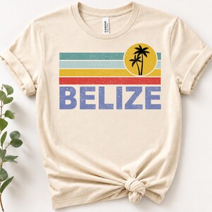 May include: A cream-colored short-sleeve t-shirt featuring a distressed graphic design. The print includes horizontal stripes in teal, light blue, yellow, and red, a yellow circle with two black palm tree silhouettes, and the word BELIZE in blue.