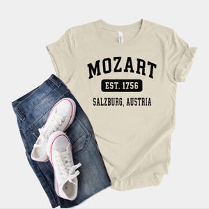 May include: A beige t-shirt with black text that reads "Mozart Est. 1756 Salzburg, Austria". The shirt is paired with blue jeans and white sneakers.