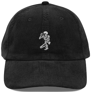 May include: A black corduroy baseball cap featuring an embroidered white graphic of a skeleton kneeling while holding a camera to its eye. The cap has a structured crown, a curved brim, and a button on top, with a classic six-panel construction.
