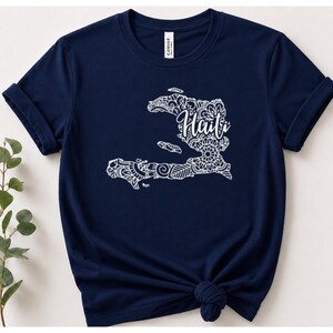 May include: A navy blue short-sleeve t-shirt featuring a white graphic of the map of Haiti filled with intricate mandala-style patterns. The word Haiti is written in a white cursive script font across the center of the map design.