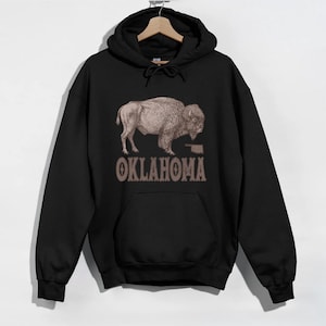 Puede incluir: Sudadera con capucha negra con una ilustración de bisonte marrón y el texto "OKLAHOMA" impreso en la parte delantera.