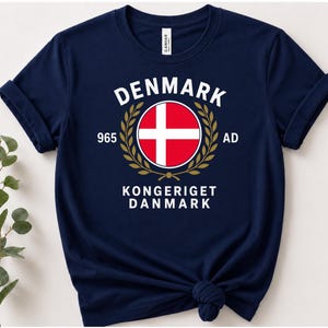 May include: A navy blue short-sleeve t-shirt featuring a central graphic with the text DENMARK, 965 AD, and KONGERIGET DANMARK. The design includes a circular Danish flag emblem surrounded by a gold laurel wreath centered on the chest.