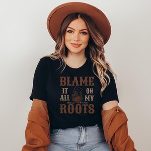 Op de afbeelding: Een zwart T-shirt met korte mouwen en de bruine tekst BLAME IT ALL ON MY ROOTS met een afbeelding van de staat New Jersey in het midden. Het shirt wordt gecombineerd met een bruine hoed met brede rand en een bruin shirt met lange mouwen.