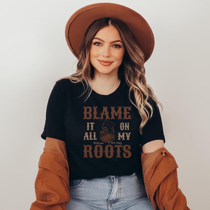 Op de afbeelding: Een zwart T-shirt met korte mouwen en de bruine tekst BLAME IT ALL ON MY ROOTS met een afbeelding van de staat New Jersey in het midden. Het shirt wordt gecombineerd met een bruine hoed met brede rand en een bruin shirt met lange mouwen.